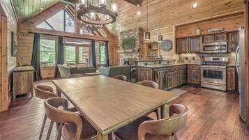 A rustic cabin interior with a wooden dining table, chairs, a stone fireplace, and a kitchen with modern appliances.