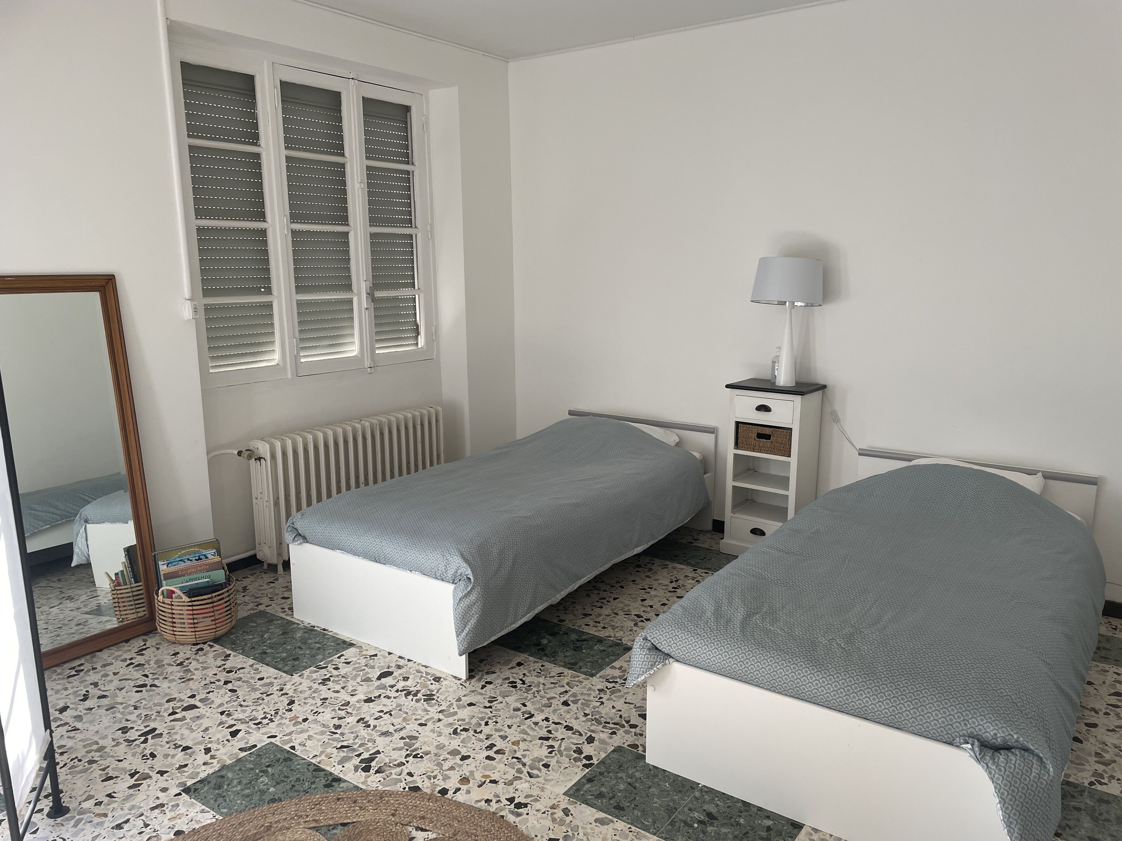Ein Schlafzimmer mit zwei Einzelbetten, einem Nachttisch mit Lampe, einem Heizkörper, einem Fenster mit Jalousien, einem Spiegel und einem Fliesenboden.
