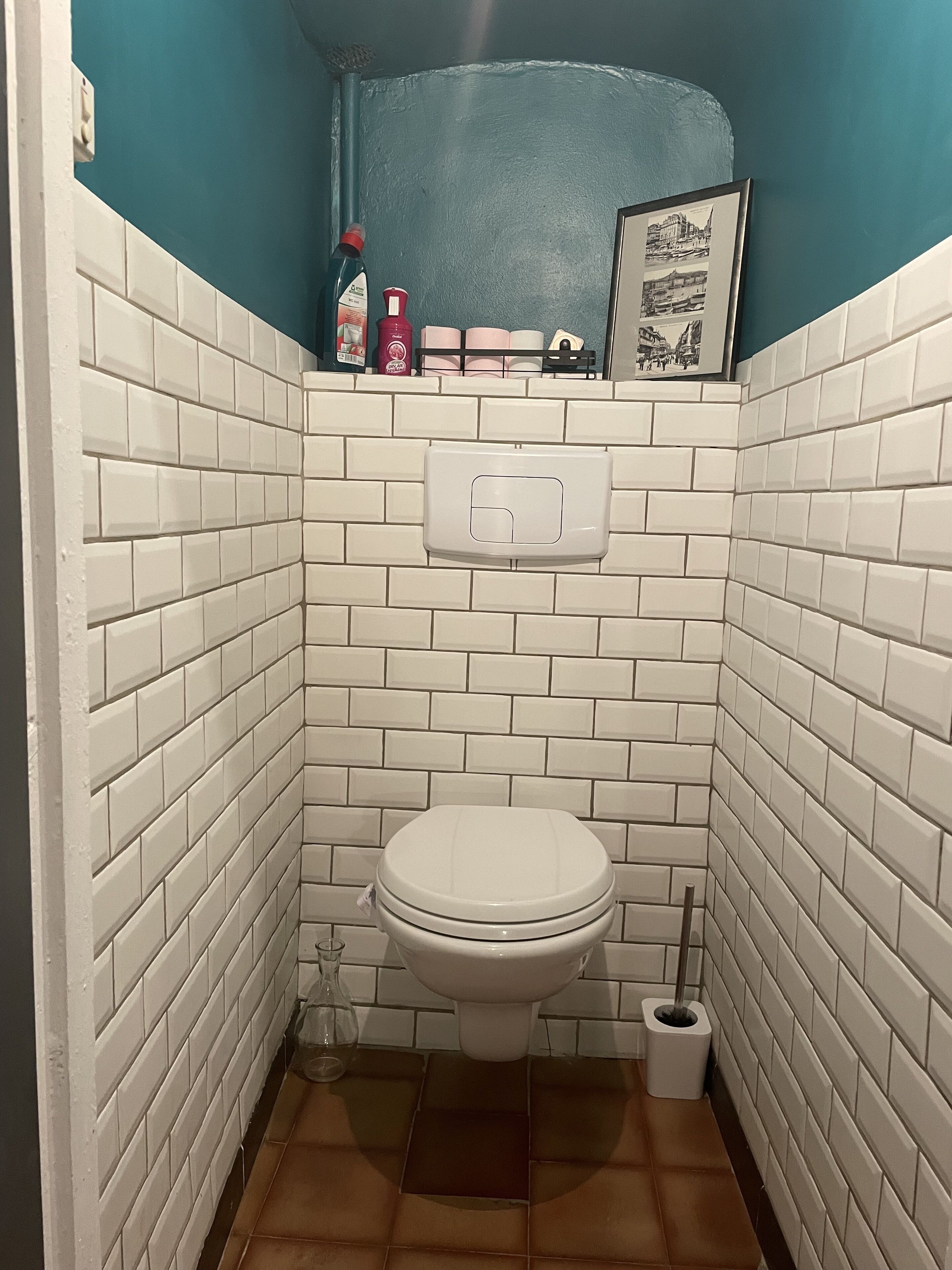 A small bathroom with white subway tiles, a white toilet, a blue wall, and a framed picture above the toilet.