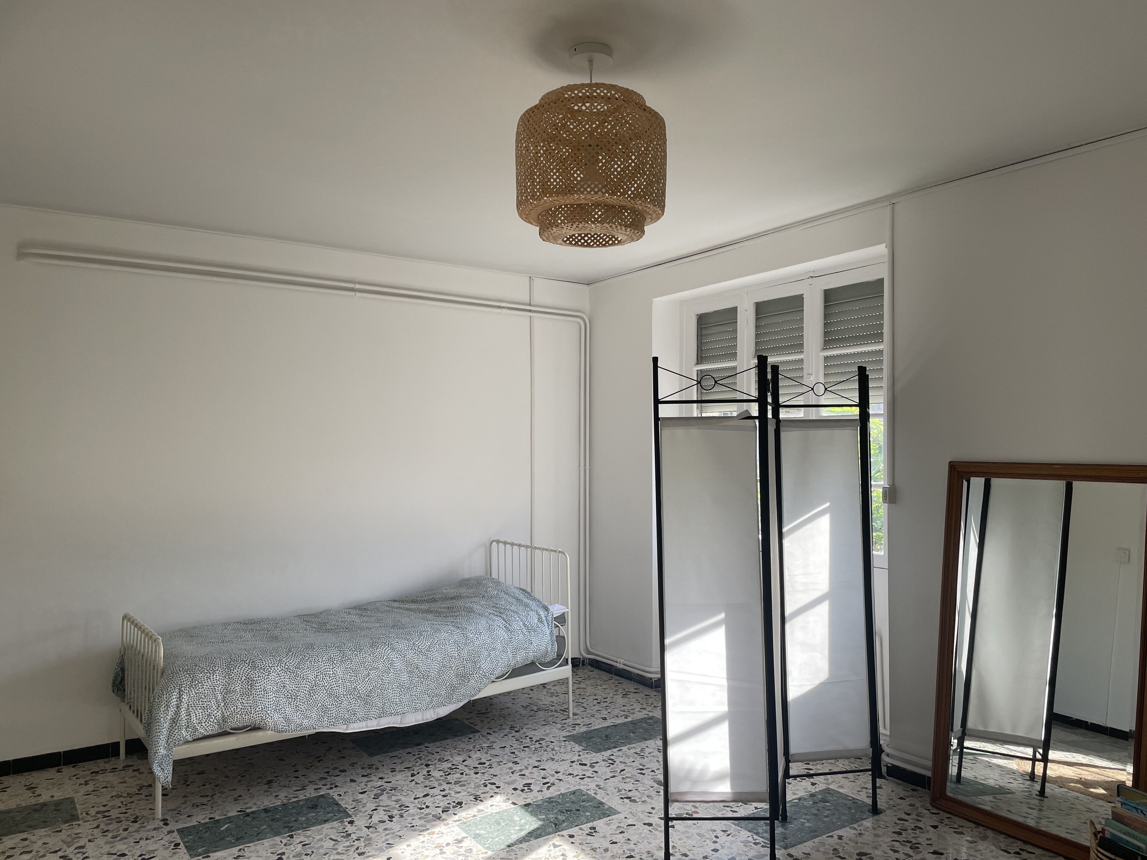 A room with a bed, a hanging light fixture, a window with blinds, and a mirror.