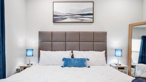 Une chambre d’hôtel avec un grand lit, deux tables de chevet avec des lampes, une tête de lit, une œuvre d’art accrochée au mur et un miroir.
