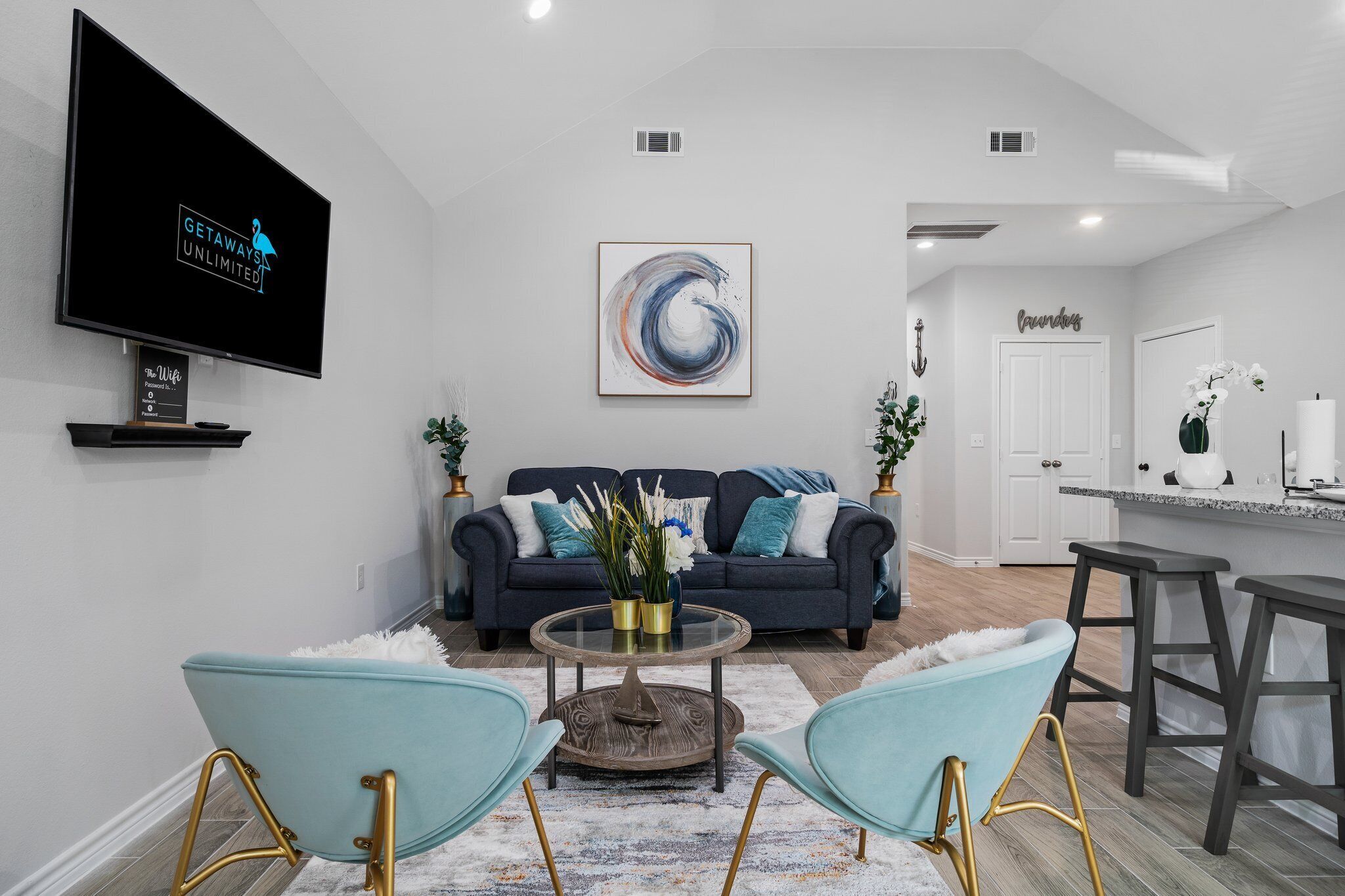 A modern living room with a dark blue sofa, two light blue chairs, a glass coffee table, and a large abstract painting.