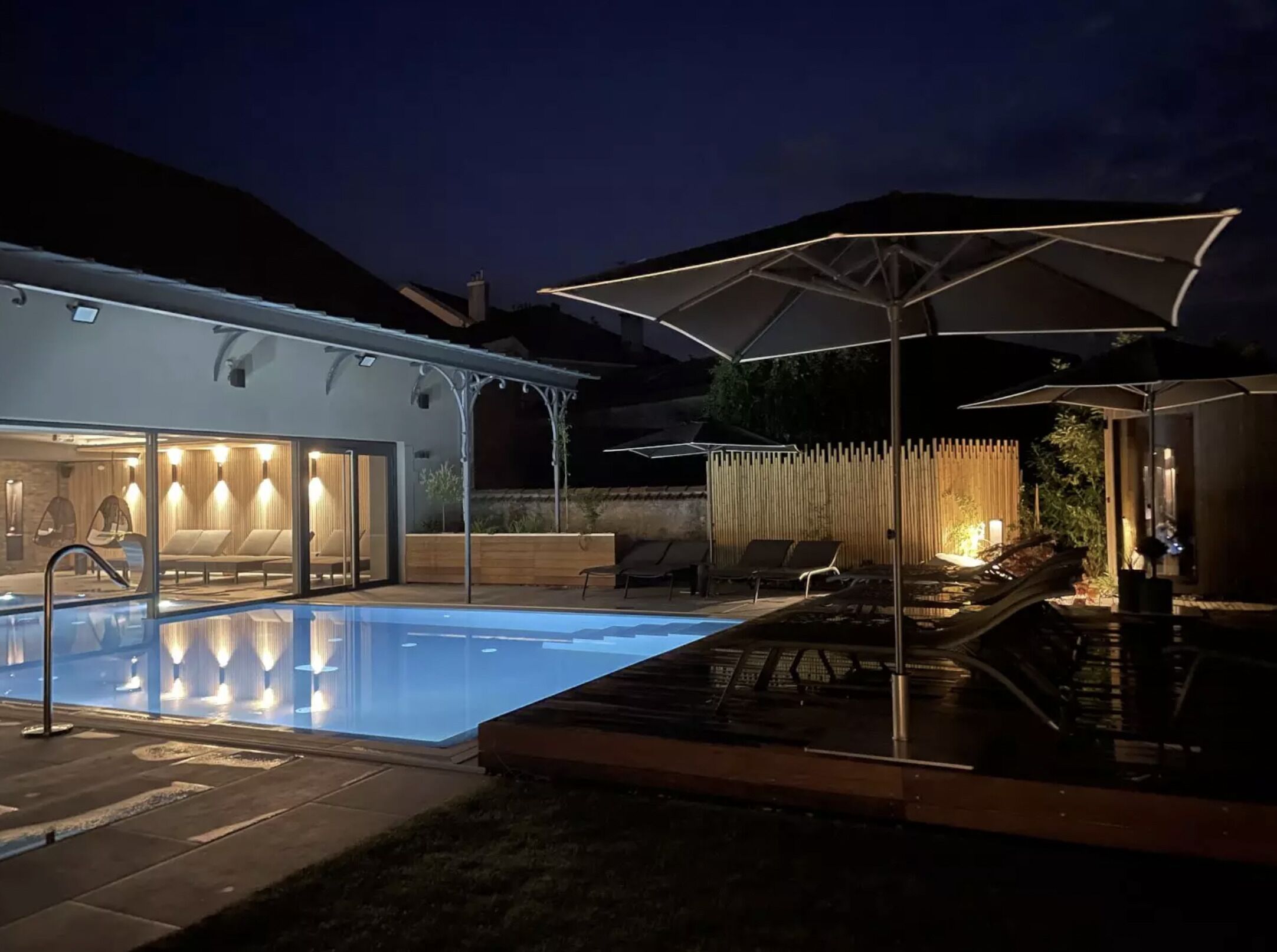 A modern outdoor pool area with lounge chairs and umbrellas, surrounded by a wooden fence and a building with glass doors.