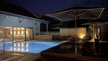 A modern outdoor pool area with lounge chairs and umbrellas, surrounded by a wooden fence and a building with glass doors.