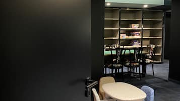 A room with a black wall and a sign reading "SALLE DE JEUX". There is a round table with chairs and a bookshelf with books and games.