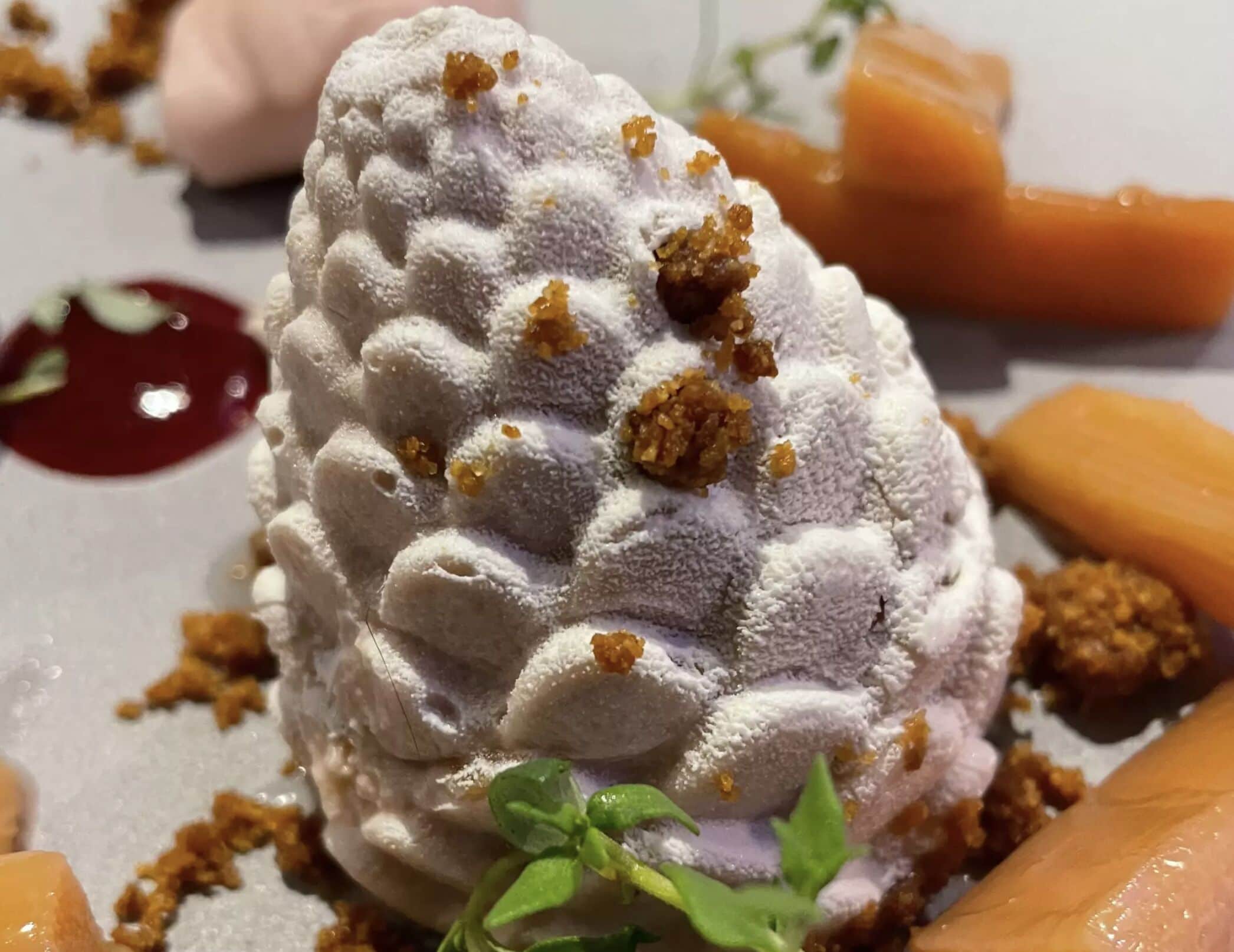 Un postre con cobertura texturizada y migas, acompañado de salsa de frutos rojos y un adorno verde.