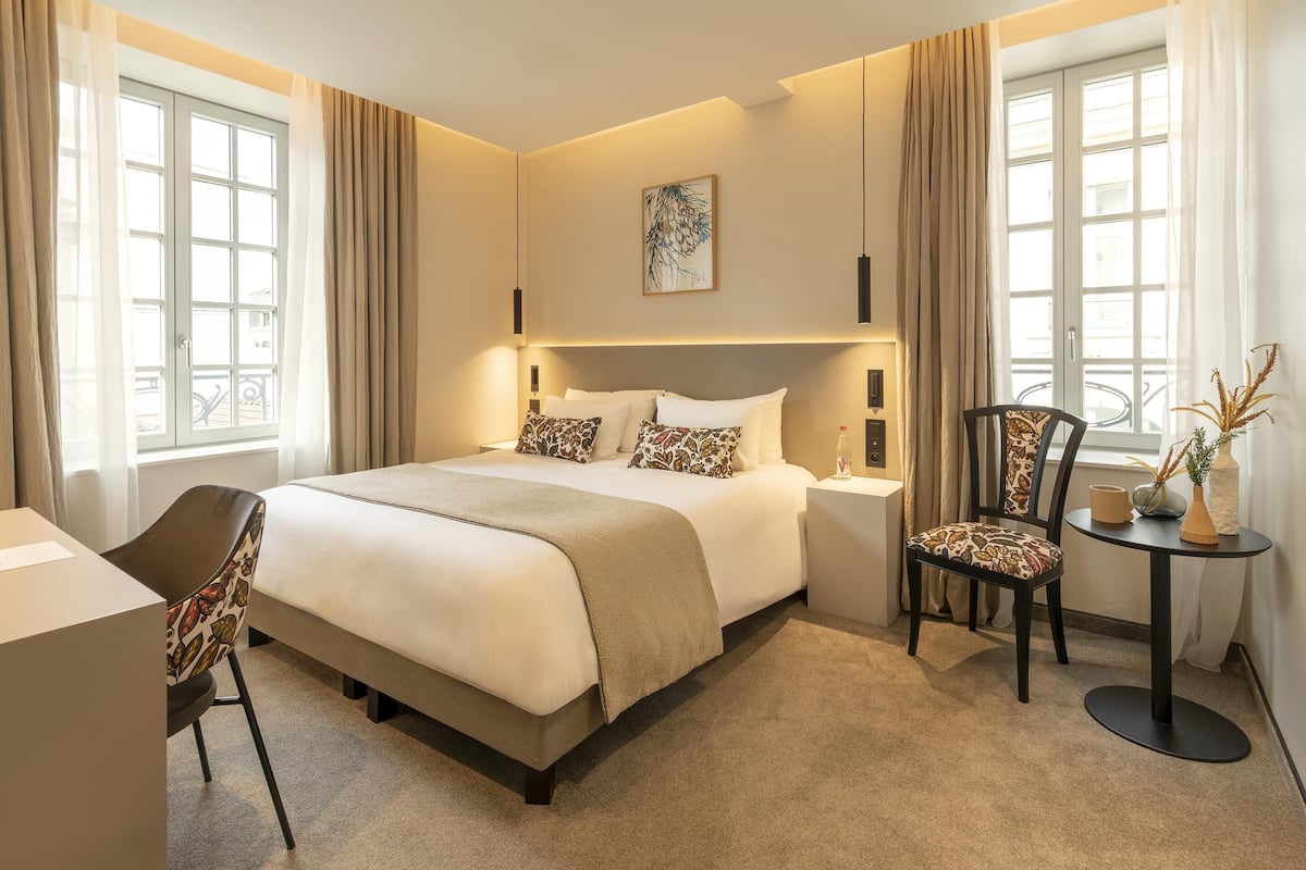 Una habitación de hotel con cama, escritorio, silla, una mesita y ventana con cortinas.