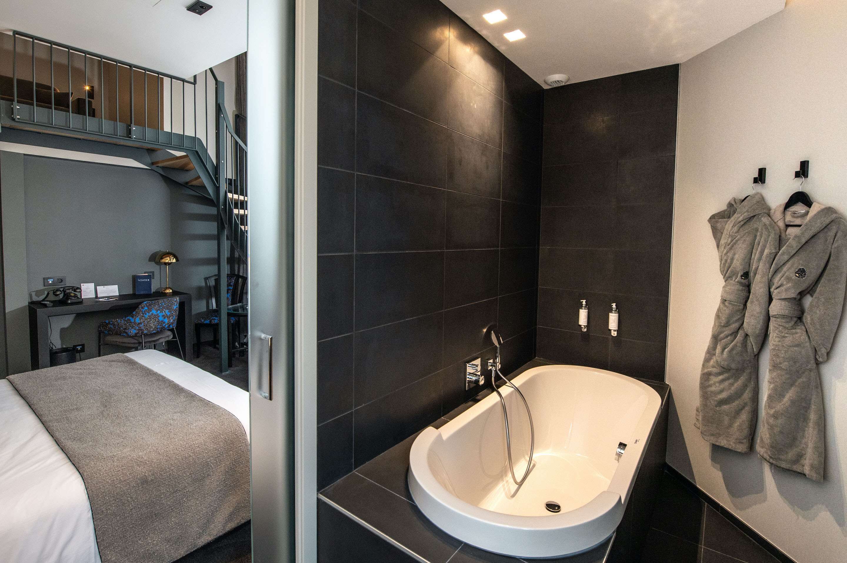 A modern hotel room with a large bed, a desk with a chair, a bathroom with a bathtub, and a staircase.