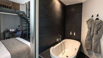 A modern hotel room with a large bed, a desk with a chair, a bathroom with a bathtub, and a staircase.