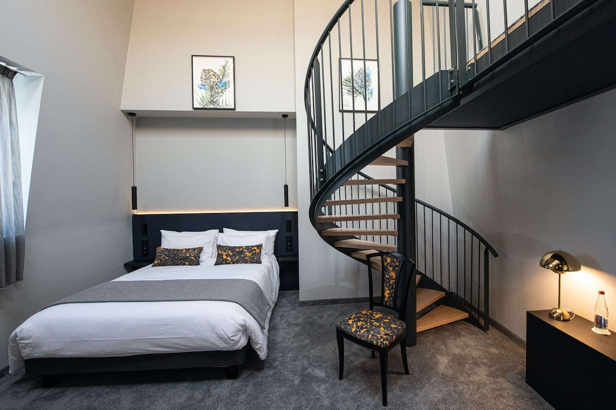 Un dormitorio con cama, una silla, una mesita de noche, una lámpara y una escalera en espiral.