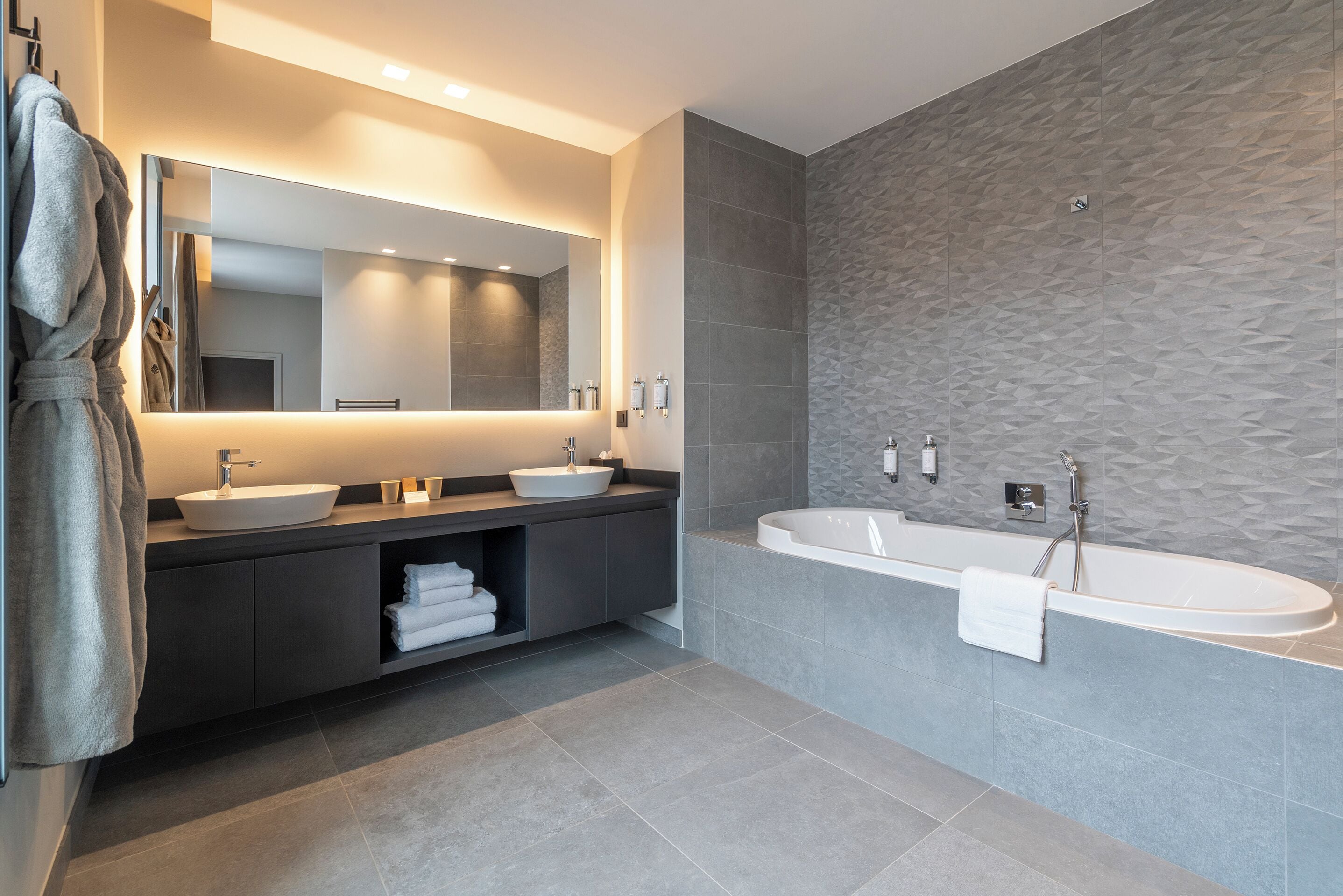 A modern bathroom with a large mirror, two sinks, a bathtub, and a towel rack.