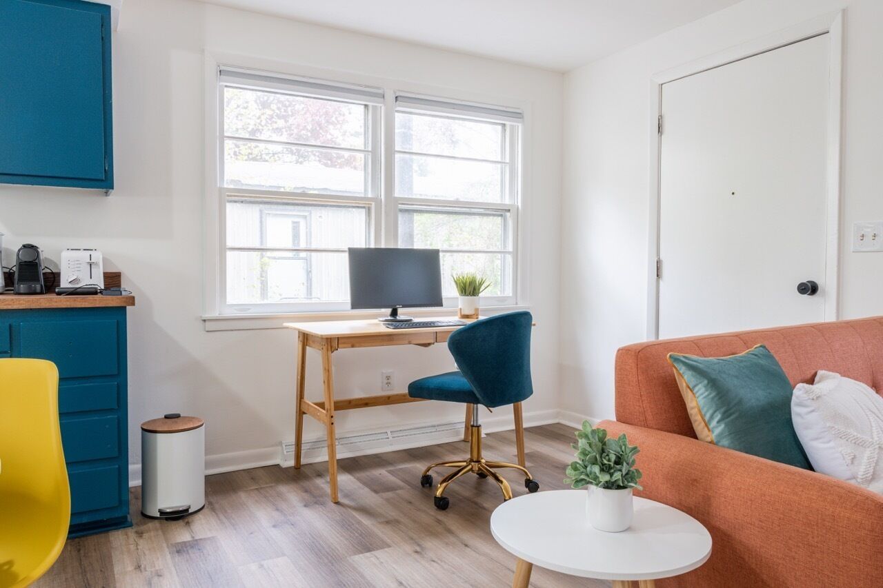En moderne stue med et træskrivebord, en computer, en blå stol, et rundt hvidt bord og en orange sofa med puder.