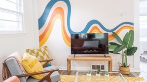 Uma sala de estar com televisão em um suporte de madeira, um mural colorido na parede, uma planta grande e uma área de estar acolhedora com almofadas.