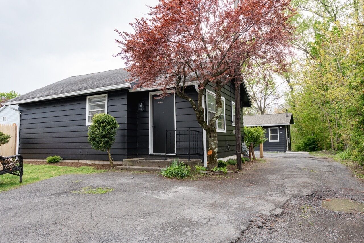 Una casa pequeña de un solo piso con fachada negra, un porche y un árbol de hojas rojas.