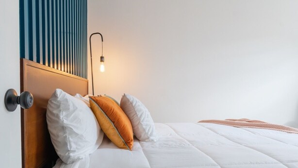 Cama bem arrumada com roupa de cama branca e alguns almofadas coloridas, cabeceira de madeira e luminária fixada na parede.