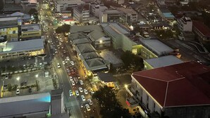 A cityscape at dusk with a sunset over the horizon, featuring a mix of high-rise buildings and smaller structures, a busy street with vehicles, and a parking area.