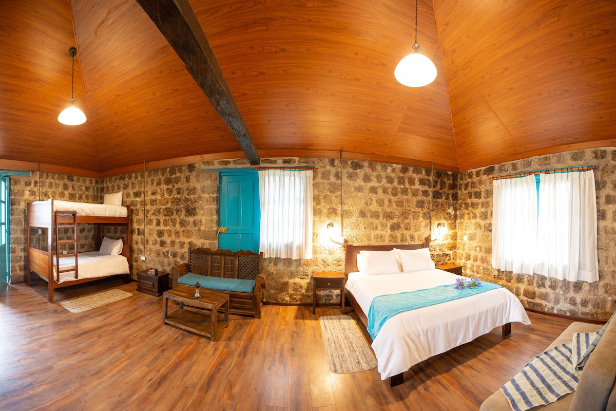 Quarto rústico de madeira com beliche, cama individual, banco de madeira, mesa pequena e paredes de pedra.