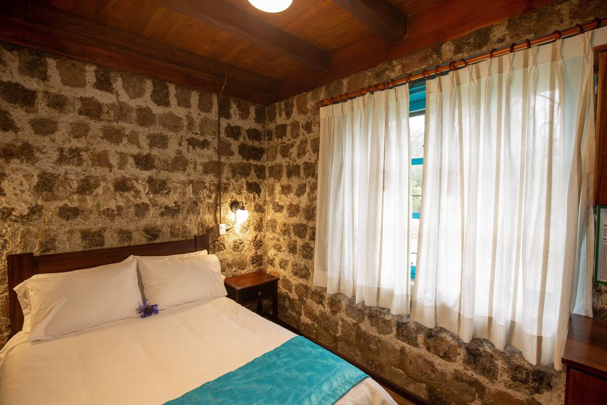 Quarto com paredes de pedra e teto de madeira, uma cama com roupa de cama branca e detalhe azul, uma mesa de cabeceira com abajur e uma janela com cortinas brancas.
