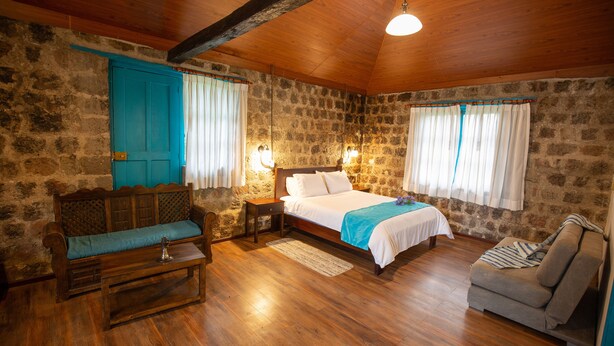 A cozy room with a wooden ceiling, stone walls, a bed with white bedding, a blue sofa, and a grey armchair. - Niebli Historical Farm and Lodge at Pululahua Volcano (Calacali)