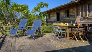 Une terrasse en bois avec des meubles d’extérieur, une table sur laquelle sont posés divers objets, et un bâtiment doté de fenêtres.