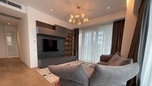 Un salon moderne avec un canapé d’angle gris, un téléviseur à écran plat fixé sur un mur foncé et un lustre.