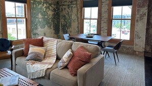 A room with a worn brick wall, a sofa with patterned cushions, a dining table with chairs, and large windows offering a view of the city. - Downtown Luxury Condo for 2! (San Antonio)