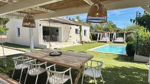 Ein Poolbereich mit Holztisch und weißen Stühlen, ein kleines Haus mit Balkon und ein Garten mit Schwimmbecken.