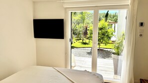 Makuuhuoneessa on sänky, seinälle asennettu televisio ja liukuovi, joka johtaa ulkotilaan, jossa on viheralue.