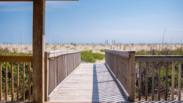 Une promenade en bois qui mène à une plage bordée de dunes de sable et dotée d’une végétation clairsemée.