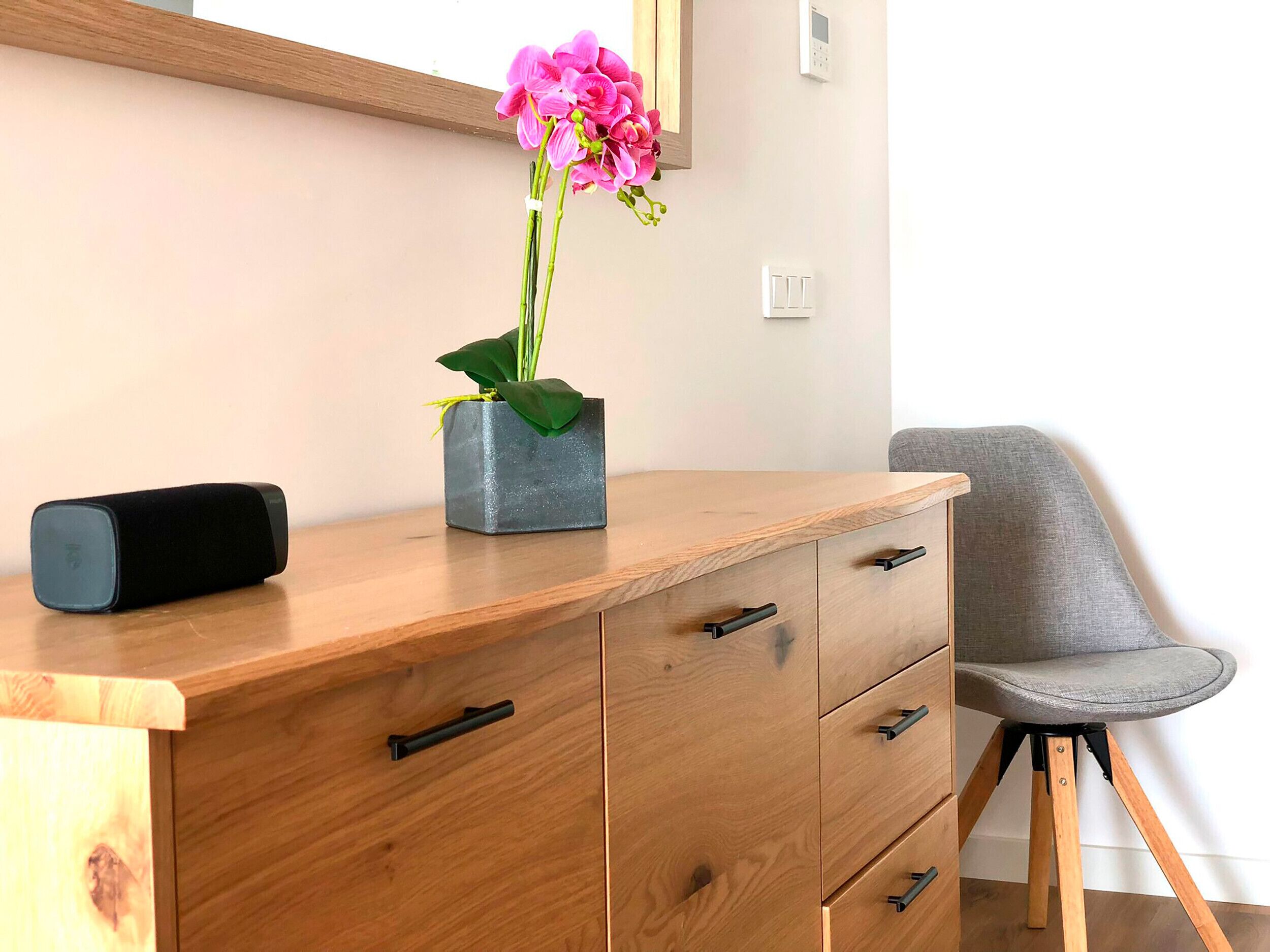 A wooden desk with a black speaker, a grey planter with a pink orchid, and a grey chair.