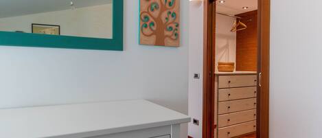 Una habitación con un aparador blanco, un espejo y una puerta de madera que da a un armario con una percha y cestas.