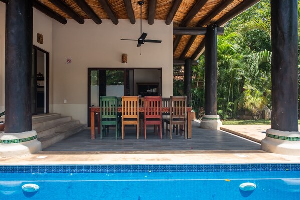 A covered outdoor area with a pool, wooden chairs, and a ceiling fan.