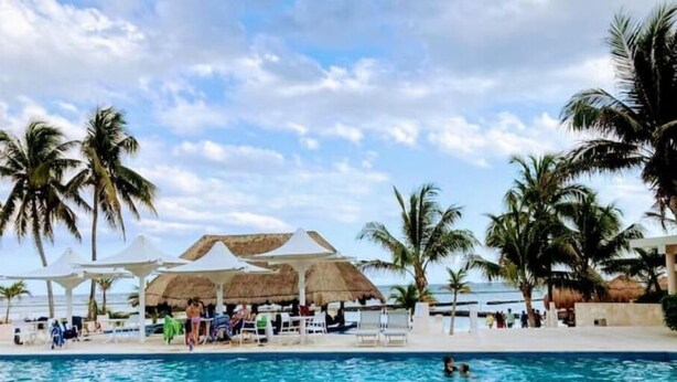 Un área de piscina con sillones de descanso y sombrillas, rodeada de palmeras y una estructura de techo de paja.