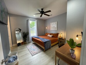 Un dormitorio con cama, ventilador de techo, ventana con cortinas, mesita de noche con lámpara, planta en maceta y alfombra.