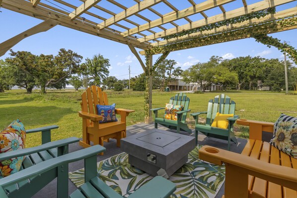 Un patio con sillas de madera y una mesa cuadrada, rodeado por una pérgola de madera.