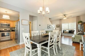 A modern kitchen with stainless steel appliances, a dining area with a dark wooden table and white chairs, and a living area with a sofa and a window.