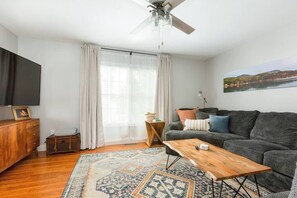A living room with a sofa, coffee table, and a television. There is a window with curtains, a ceiling fan, and a painting on the wall.