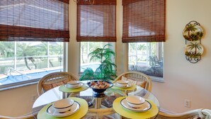A dining area with a round table set for four, surrounded by wicker chairs. There are three windows with brown blinds, a potted plant in the center, and a decorative wall piece above the table. - Near Sand & Sun, Cape Coral Home w/ Private Pool (Cape Coral)