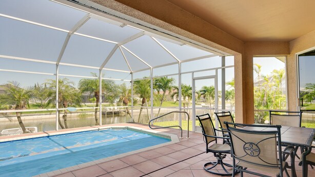 A covered outdoor pool area with a table and chairs, surrounded by glass panels and palm trees. - Near Sand & Sun, Cape Coral Home w/ Private Pool (Cape Coral)