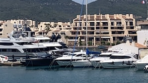 A marina with several yachts docked, a building with multiple balconies, and a mountain in the background.