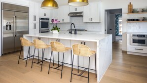 A modern kitchen with white cabinetry, stainless steel appliances, a marble island with bar stools, and gold pendant lighting. - Modern, Spacious Pool Home in Plano (Plano)