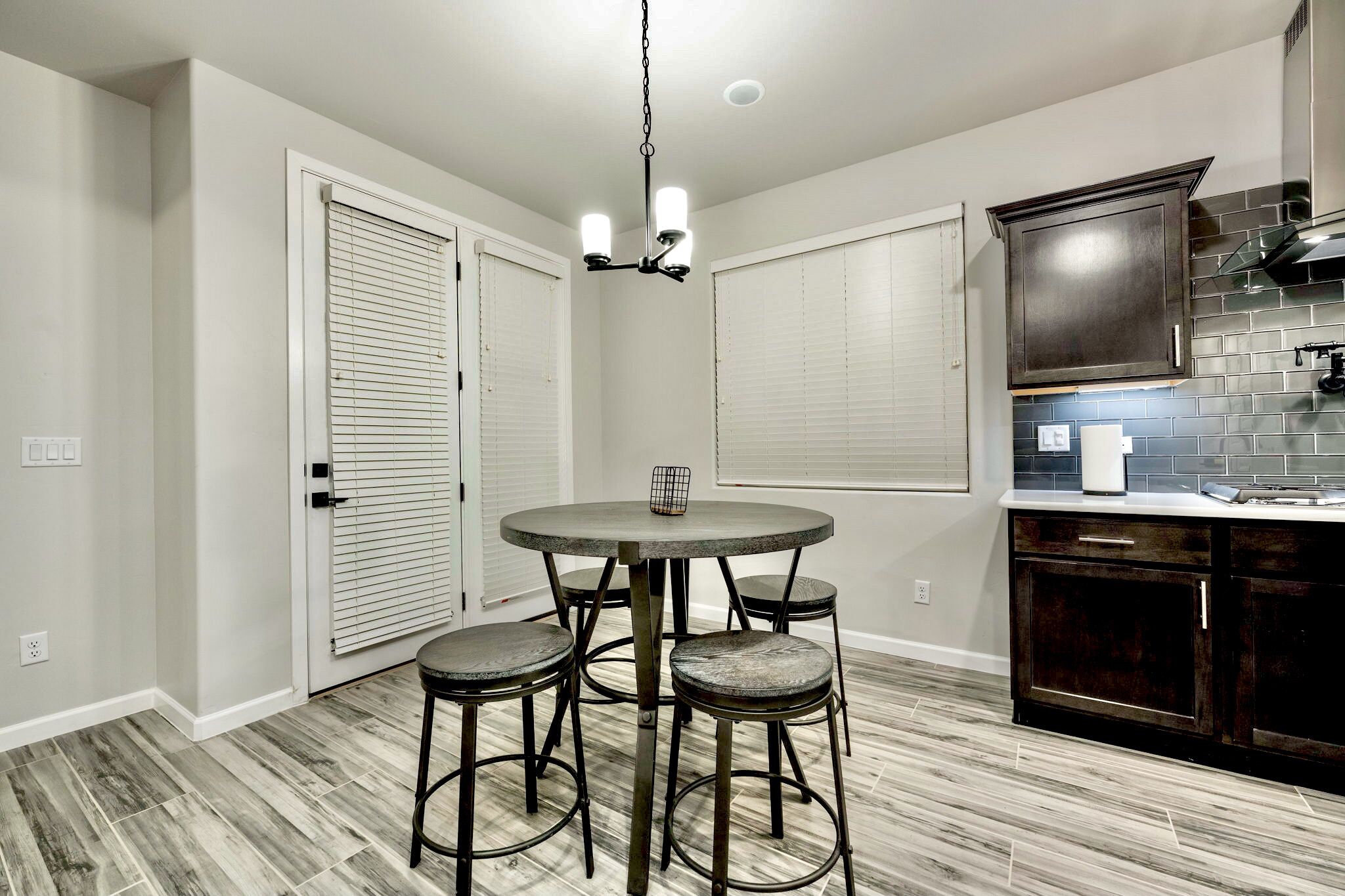 A kitchen with a round table and four stools, a dark wooden cabinet, and a light-colored tiled backsplash.