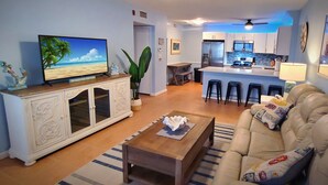 A living room with a TV, sofa, coffee table, and a kitchen in the background. - Cozy Condo Right on the Beach! (Cocoa Beach)