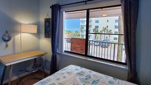 A room with a bed, a desk, a window with a view of a parking area and buildings. - Cozy Condo Right on the Beach! (Cocoa Beach)