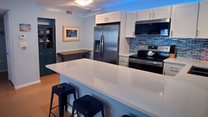 A modern kitchen with white cabinets, a stainless steel refrigerator, a microwave, and a large island with bar stools. - Cozy Condo Right on the Beach! (Cocoa Beach)