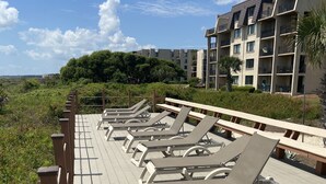 Een terras met ligbedden, een houten leuning en uitzicht op een meerlaags gebouw met balkons.