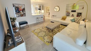 A modern living room with a white sofa, a coffee table, a dining area with a kitchen in the background, and a large mirror on the wall. - Mid Century Spacious House 4BR/Heated pool (Oakland Park)