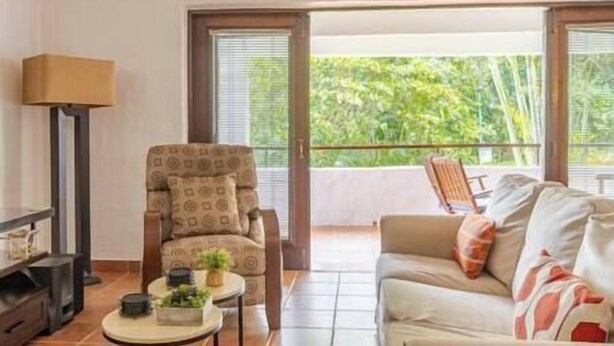 A living room with a sofa, a patterned armchair, a small table with a lamp, and a view of greenery through large windows. - Cozy Golf Course View Villa inside the Wyndham Grand Rio Mar Resort (Rio Grande)