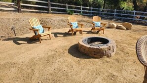A sandy outdoor area with three wooden Adirondack chairs with blue cushions, a stone fire pit, and a white fence in the background. - Casita dos Robles Fallbrook's Favorite Dog-friendly Country Escape (Fallbrook)