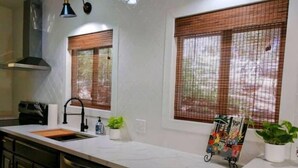 A kitchen with a white countertop, black cabinets, a stainless steel sink, and a black faucet. There are two windows with brown blinds, a small potted plant, and a magazine rack with books. - Casita dos Robles Fallbrook's Favorite Dog-friendly Country Escape (Fallbrook)